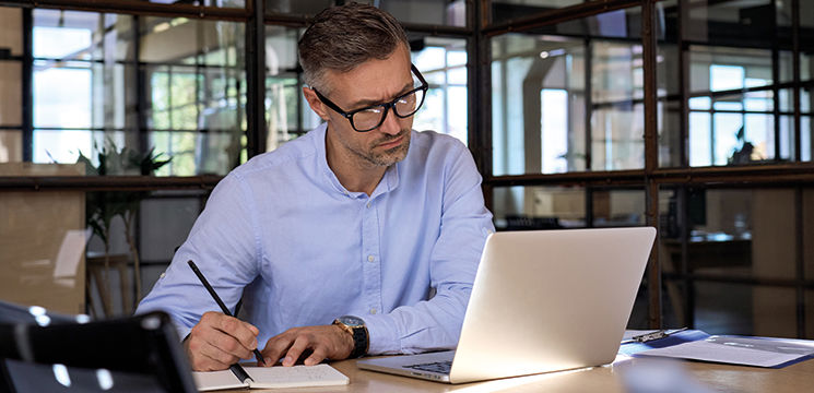 Professional man at a desk writing in a note pad while looking at a laptop screen